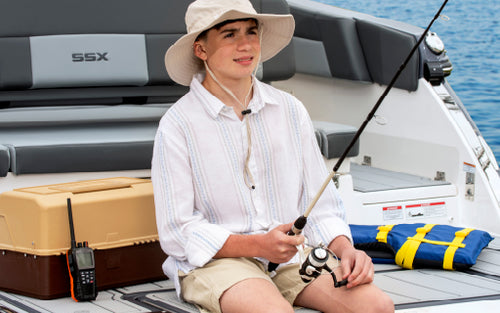 A kid fishing off the back of a boat with a tackle box and marine radio next to him.