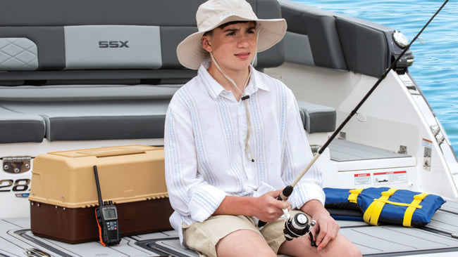 A kid fishing off the back of a boat with a tackle box and marine radio next to him.