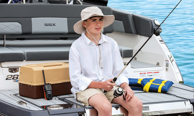 A kid fishing off the back of a boat with a tackle box and marine radio next to him.